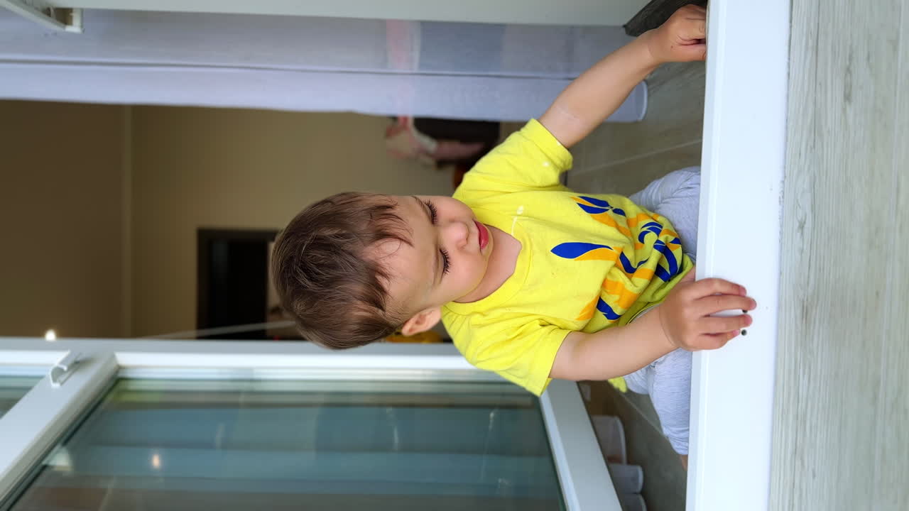 Lovely little boy sitting at the door to a balcony. Cute child wearing yellow t-shirt at home. Vertical video.