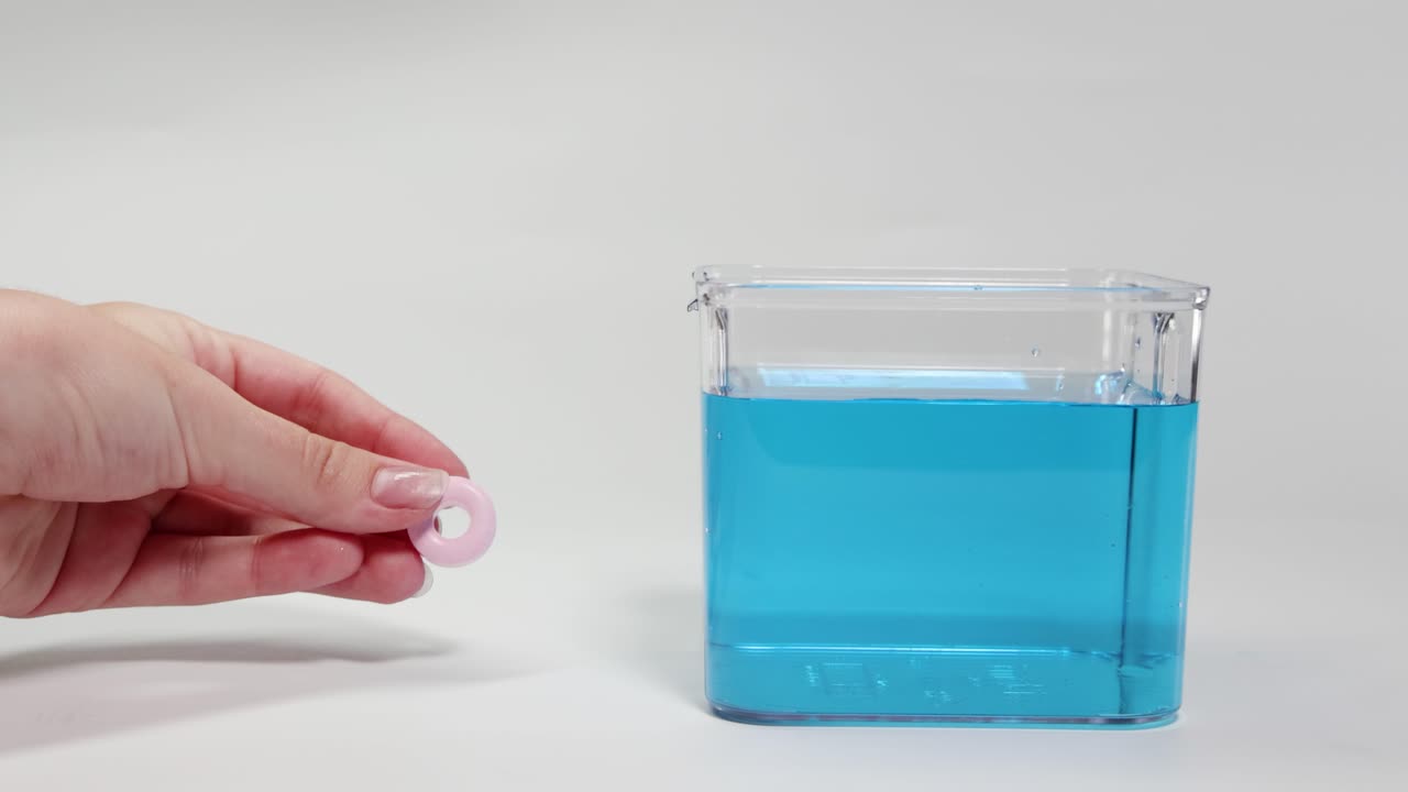 A hand drops a candy into a blue liquid, observing its buoyancy. Bright lighting highlights the clear container and vibrant colors