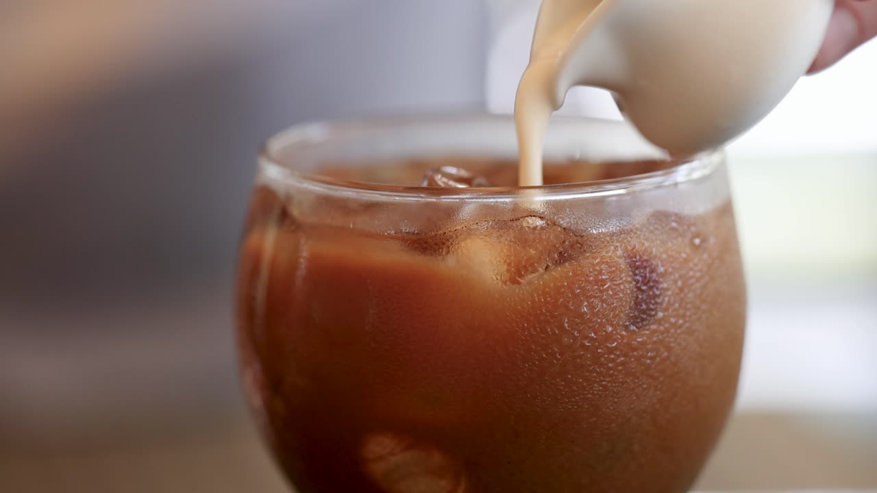 Il latte viene versato nel caffè freddo, creando motivi a spirale. Il video cattura la miscela rinfrescante in un bicchiere trasparente.
