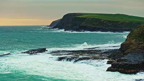 Green-orange-glow-spreads-across-horizon-as-ocean-waves-crash-at-base-of-cliffs-in-New-Zealand