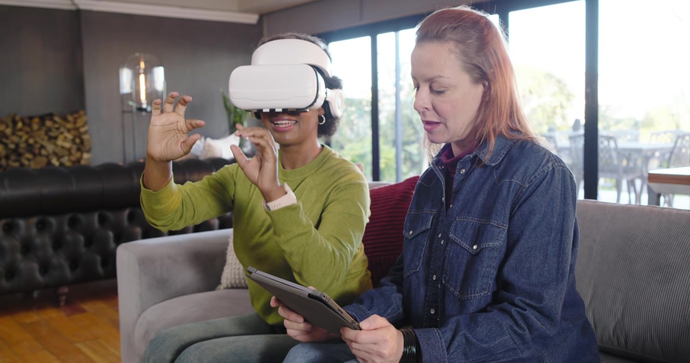 Wanita beragam memandu dengan tablet, pemakai headset VR menjelajahi, meraih, mencubit di sofa ruang tamu