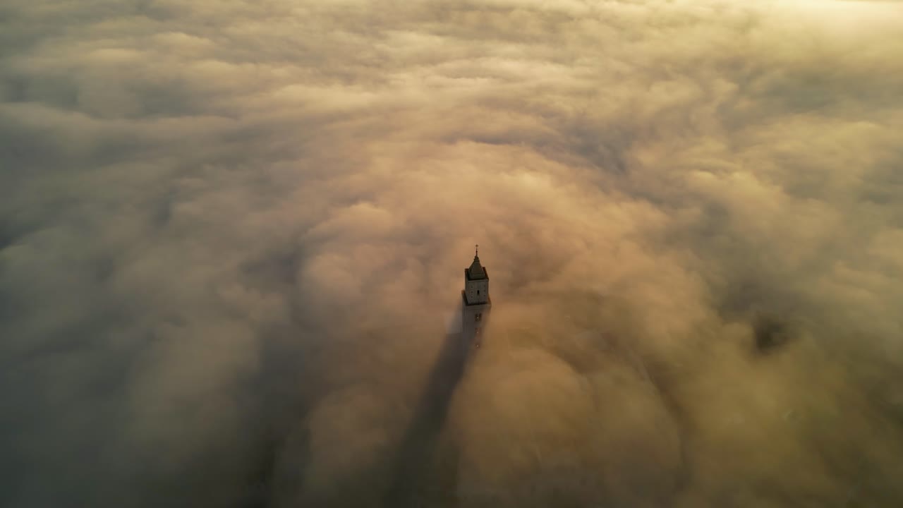Epic view if a church tower protruding through low hanging clouds during sunrise 4k drone shot video. Drone is slowly flying above a church tower that is only partially visible, sunrise, golden hour