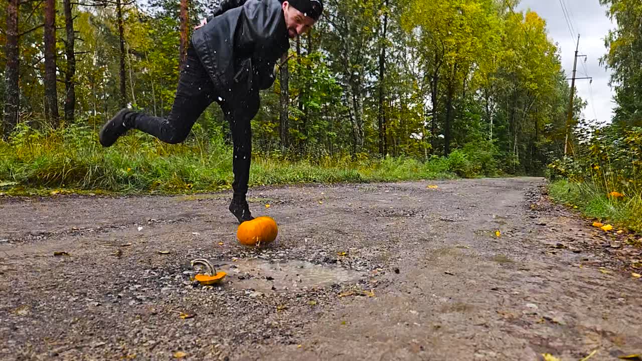 Un uomo che lancia e distrugge una zucca di Halloween arancione al rallentatore in una pozzanghera su una strada sterrata in un luogo rurale. L'uomo indossa abiti neri e una giacca di pelle durante l'autunno, a ottobre.
