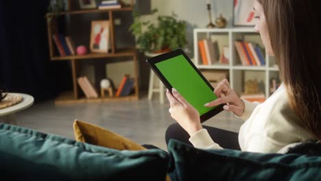 woman using gadget tablet with green screen. unrecognizable female person swiping on chroma key, sitting on sofa in living room. online shopping, surfing internet. spare time at home, leisure