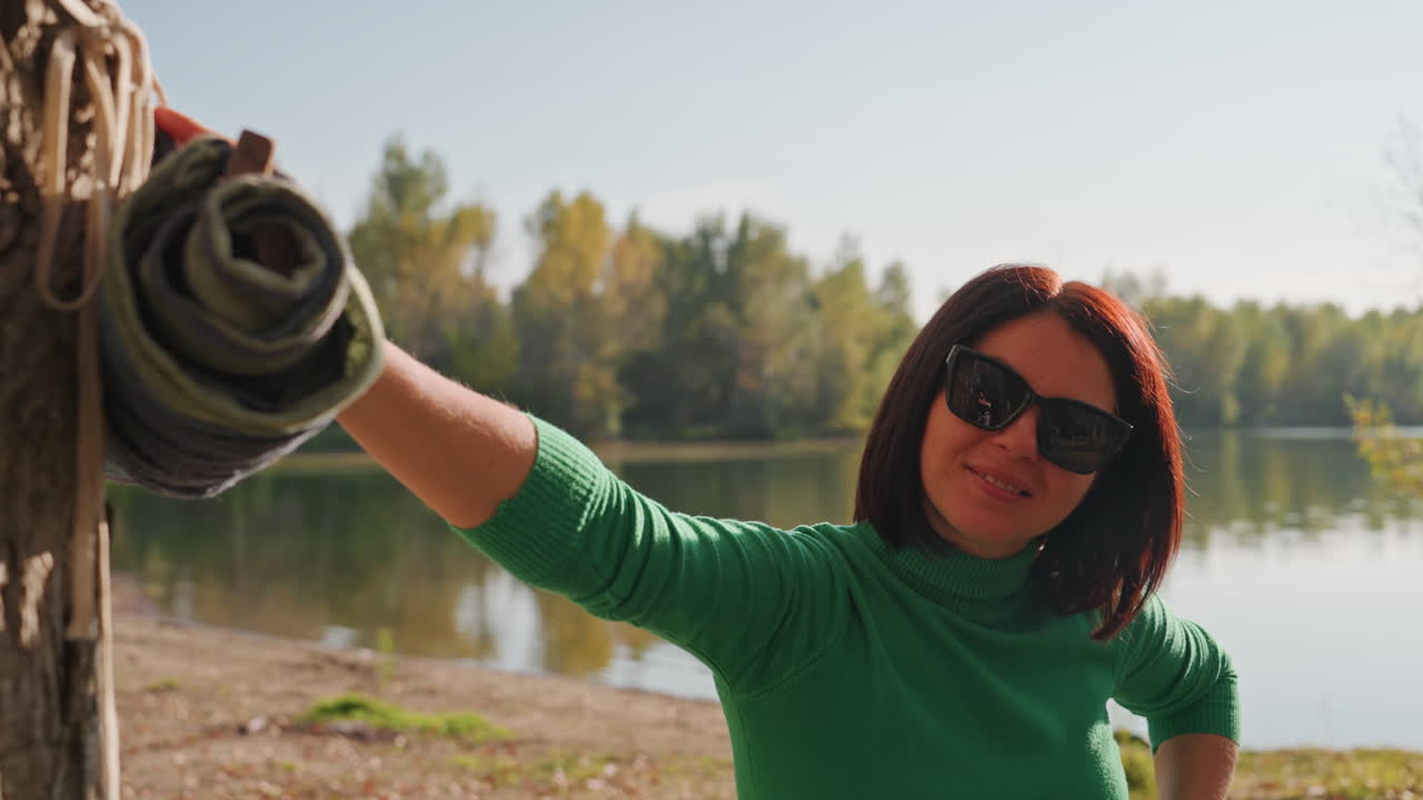 Donna che tiene una coperta arrotolata in riva al lago, sorridente e che offre attrezzatura con un gesto di benvenuto, sole sull'acqua, sfondo di alberi, atmosfera amichevole da host del campeggio e ospitalità rilassata all'aperto per gli ospiti.