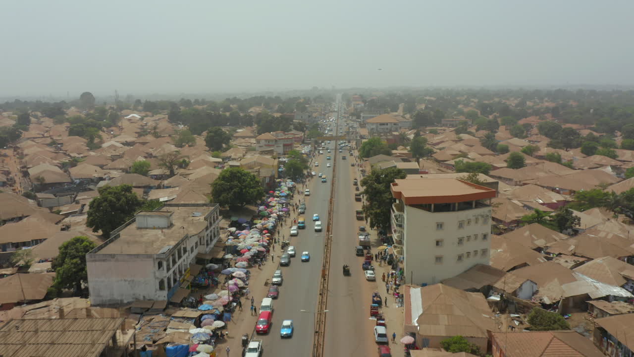 The busy main avenue and adjacent street market in Bissau, Guinea-Bissau. Shows heavy traffic, commercial activity, and the endless spread of the capital's low-rise urban density