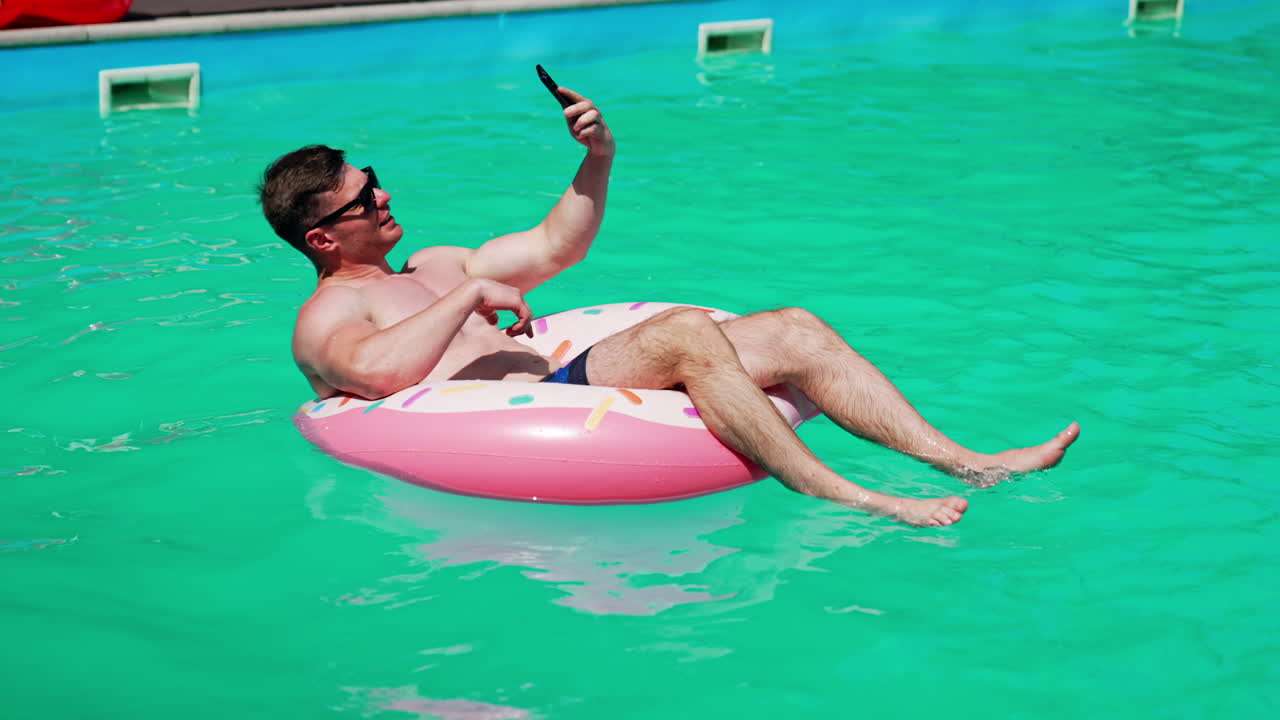 Relaxed Caucasian sportive man swimming in the pool. Man sits in the inflatable donut having a video chat on his phone.
