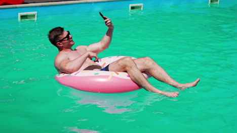 relaxed caucasian sportive man swimming in the pool. man sits in the inflatable donut having a video chat on his phone.