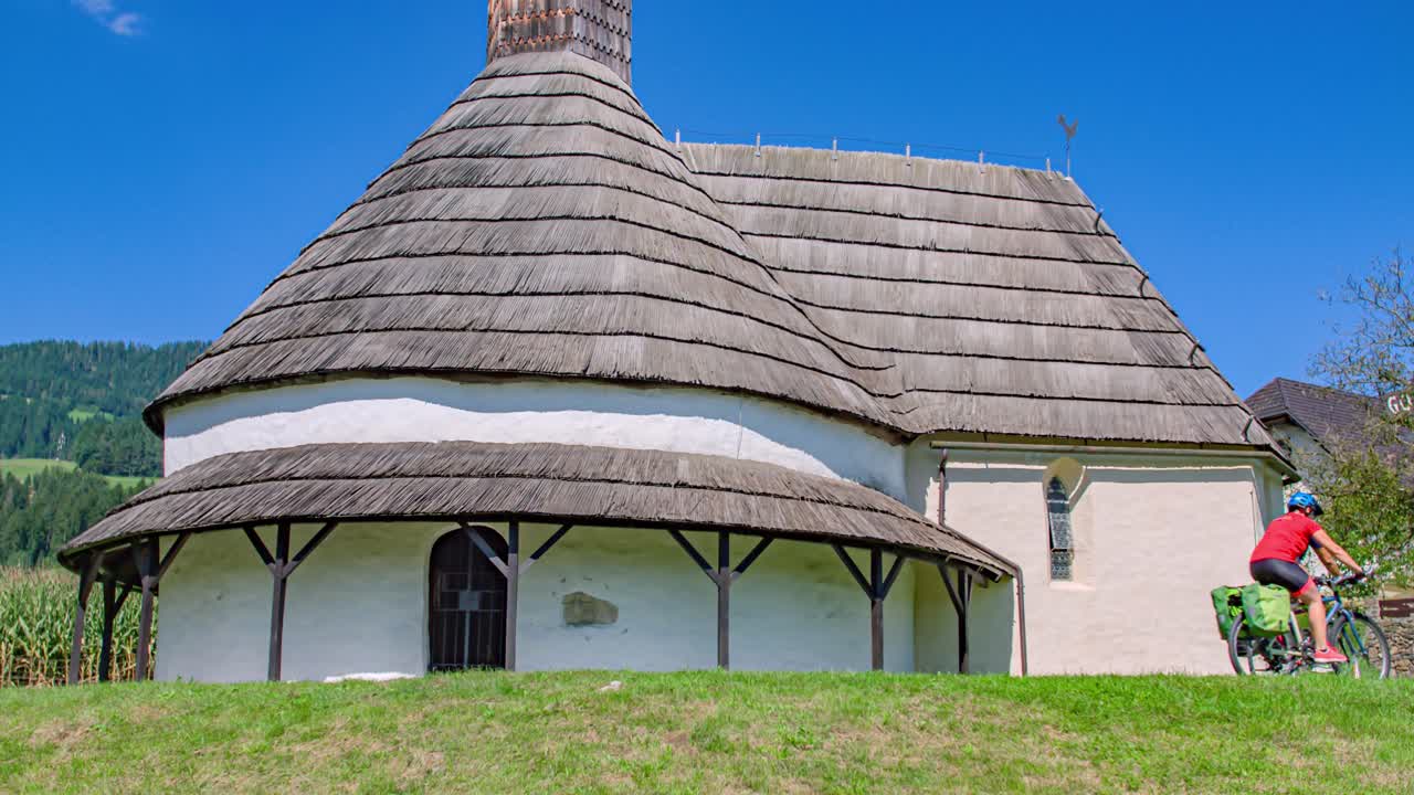 Tilting shot revealing two bikers riding beside Rotunda of Saint John the Baptist in Muta, Slovenia
