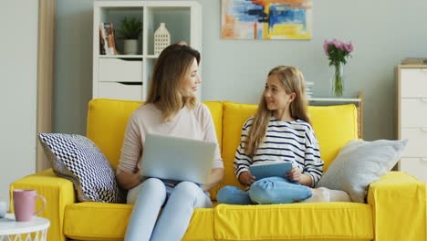 blonde mother and daughter sitting on yellow couch while they using laptop computer and tablet and talking in the living room 1