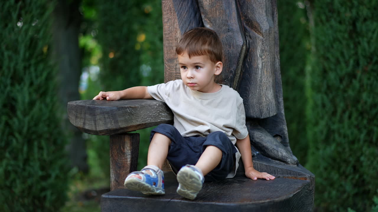 Kid climbs on the chair near the wooden sculpture. Little toddler boy spending time outdoors in summer.