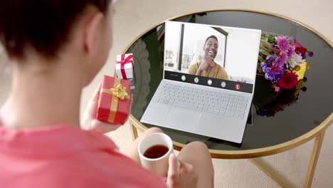 Caucasian-woman-holding-red-gift-using-laptop-with-african-american-man-on-screen