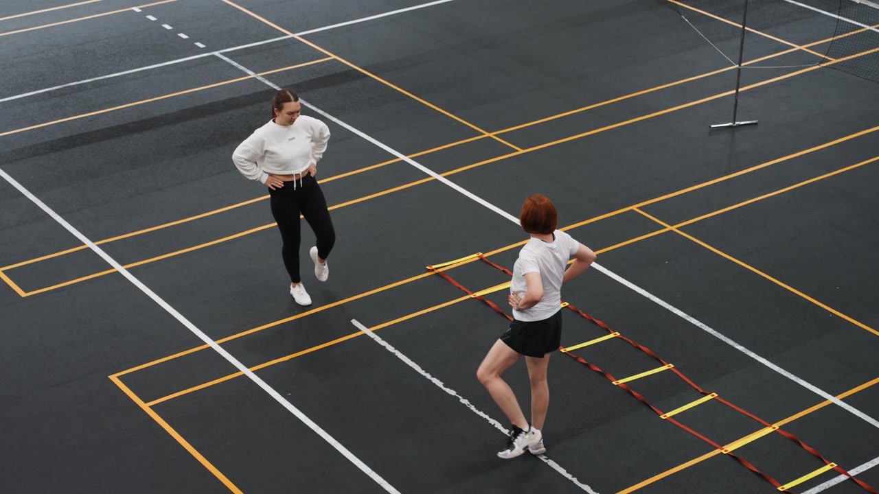Pareja de bádminton calentando con escalera de agilidad, dos jugadores replicando elevaciones rápidas de rodilla y pasos laterales en una cancha cubierta marcada cerca del poste de la red, preparación de alta energía y enfoque en el partido.
