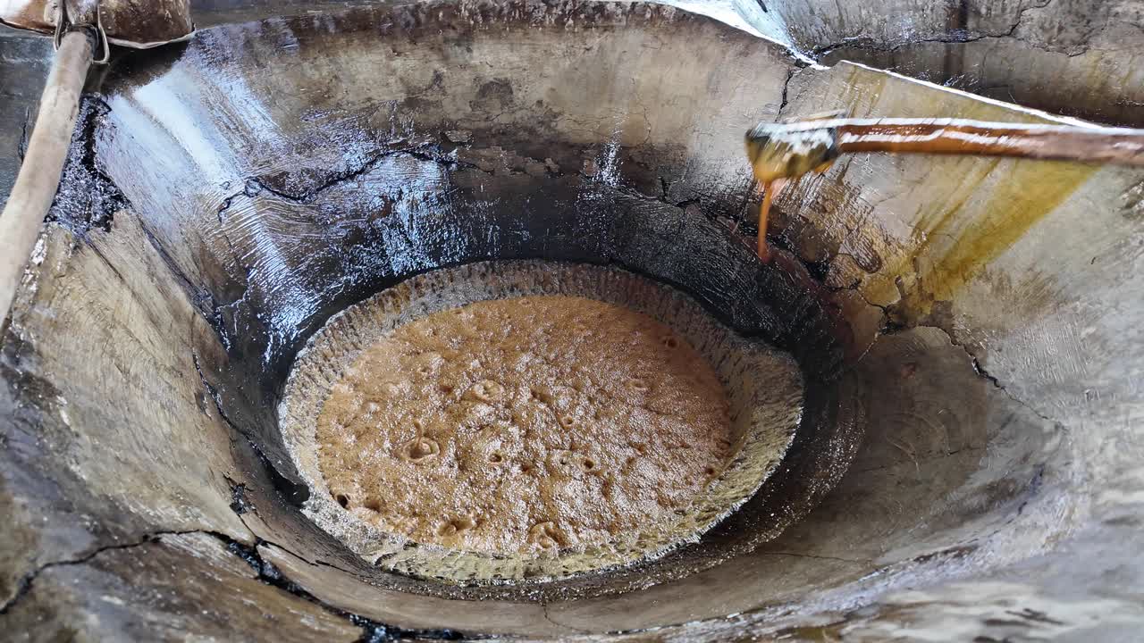 Man stirs thick, bubbling caramel sugar in a rustic vat over open heat source
