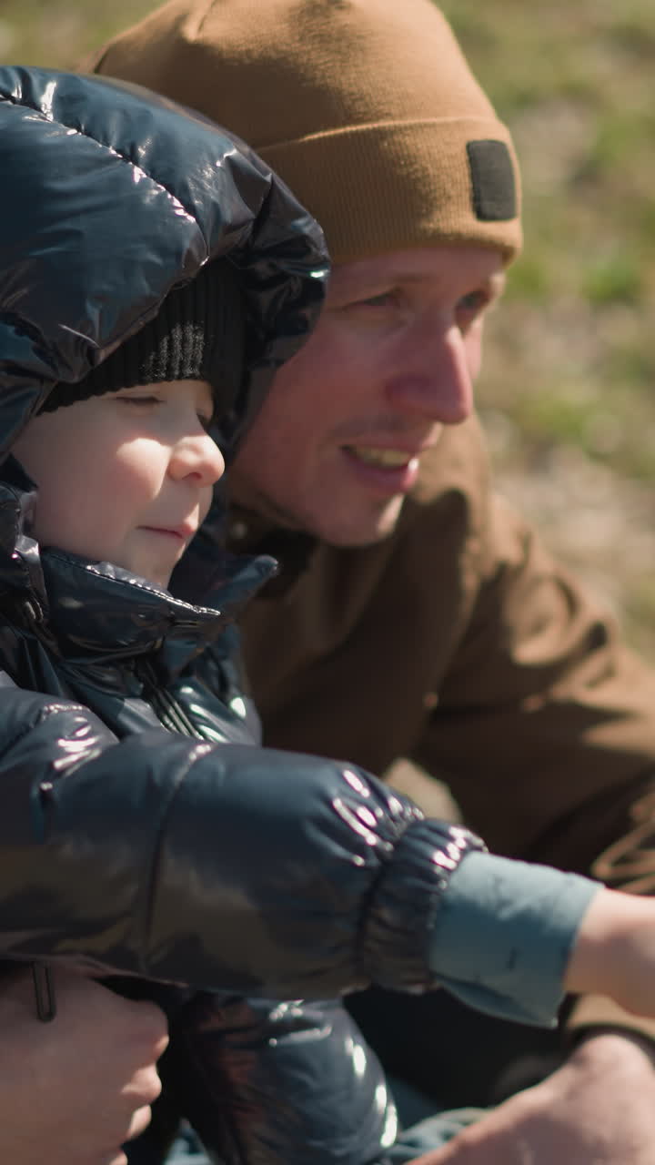 A close-up of a father squatting and carrying his son on his leg, the child, dressed in a shiny black jacket, is pointing at something and speaking with excitement