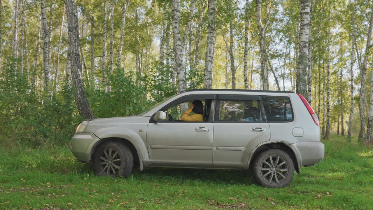 SUV dirigindo por bosque de bétulas com faróis acesos, motorista mulher visível dentro se preparando para sair do acampamento, close-up de casca de bétula e rodas enlameadas, luz do entardecer e movimento criam exploração com atmosfera envolvente