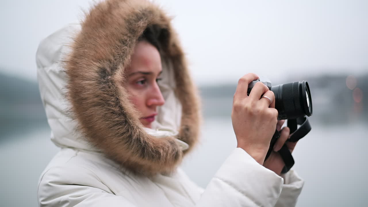 A woman dressed in a warm coat takes photos near a lake, appreciating the serene environment on a cool, overcast day. She focuses on capturing the moment