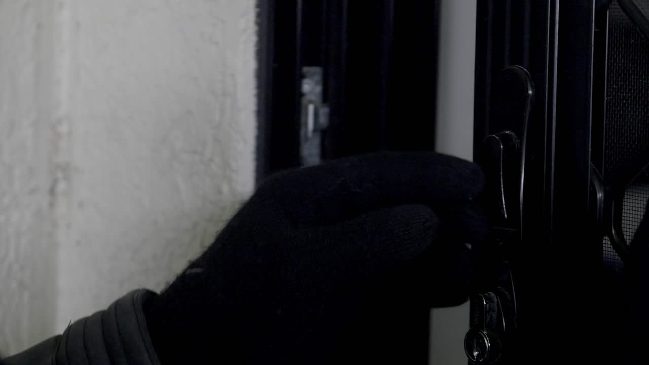 un ladrón con guantes negros, abre silenciosamente una puerta de pantalla de seguridad para entrar en una propiedad