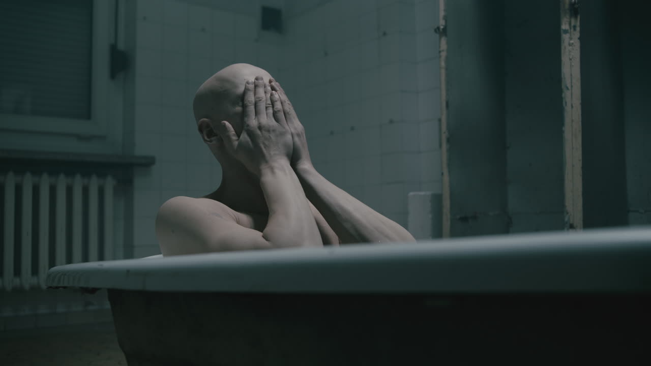 Pale bald skinny man takes a bath in the creepy old bathroom at night