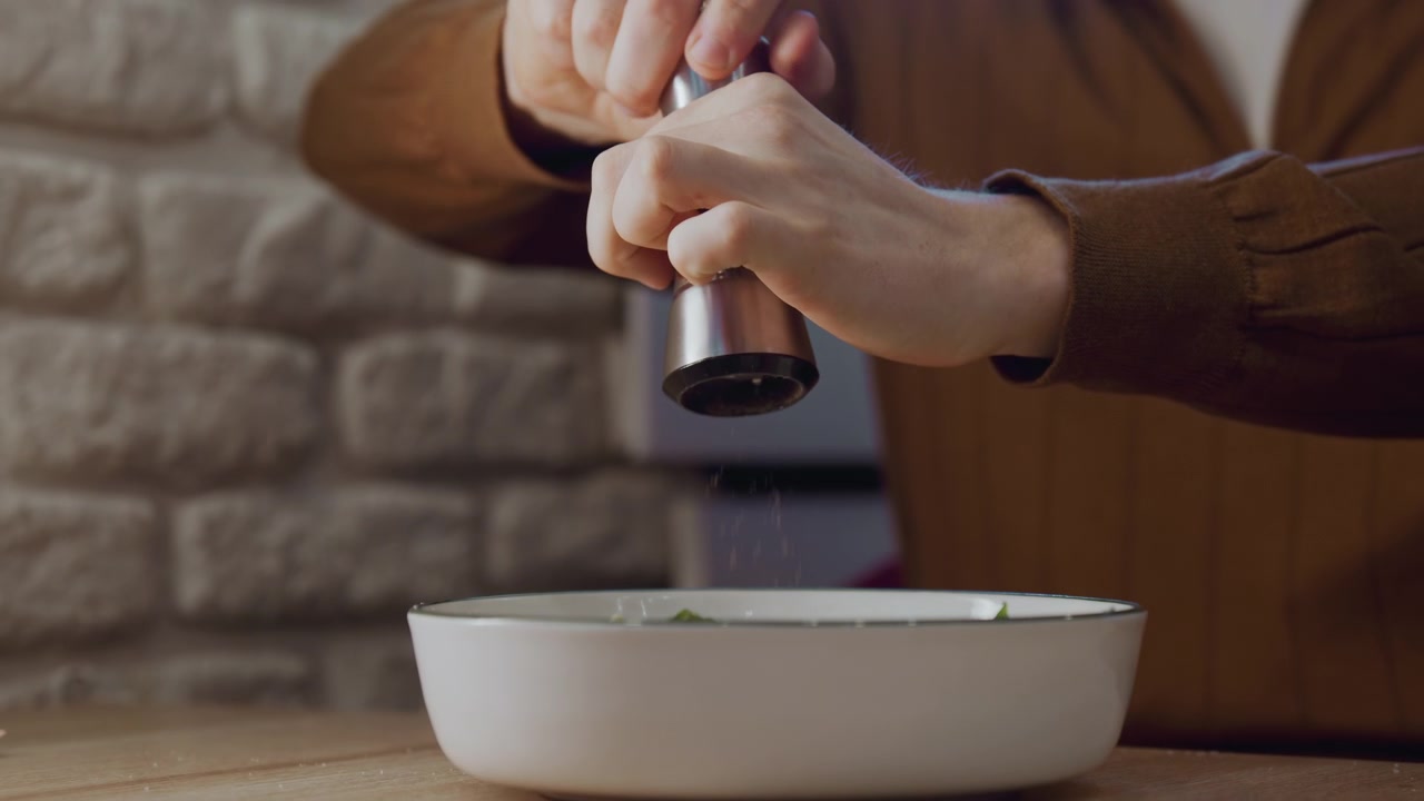 Primo piano di una persona che condisce il cibo con il pepe in cucina. Mani che tengono un macinino di metallo sopra una ciotola bianca su un tavolo di legno con sfondo di parete in pietra. Azione di preparazione del cibo visibile