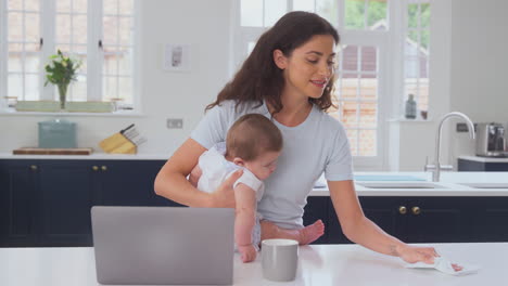 mother with baby son working from home on laptop in kitchen and cleaning surface