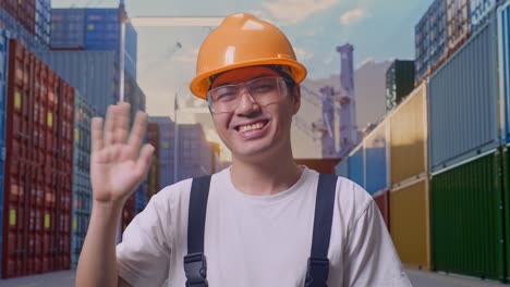 primer plano de un trabajador asiático con gafas de protección y casco de seguridad sonriendo, agitando la mano y diciendo adiós mientras estaba de pie en el almacén del patio de contenedores