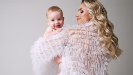 caucasian woman with long wavy blonde hair turning around with baby in hands. mother waving her cute child. white backdrop.