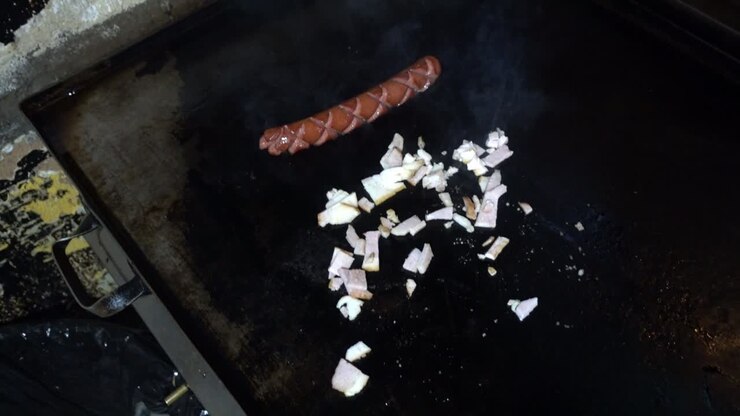 Primo piano di uno chef che cucina cipolle e un hot dog su una griglia in una cucina.