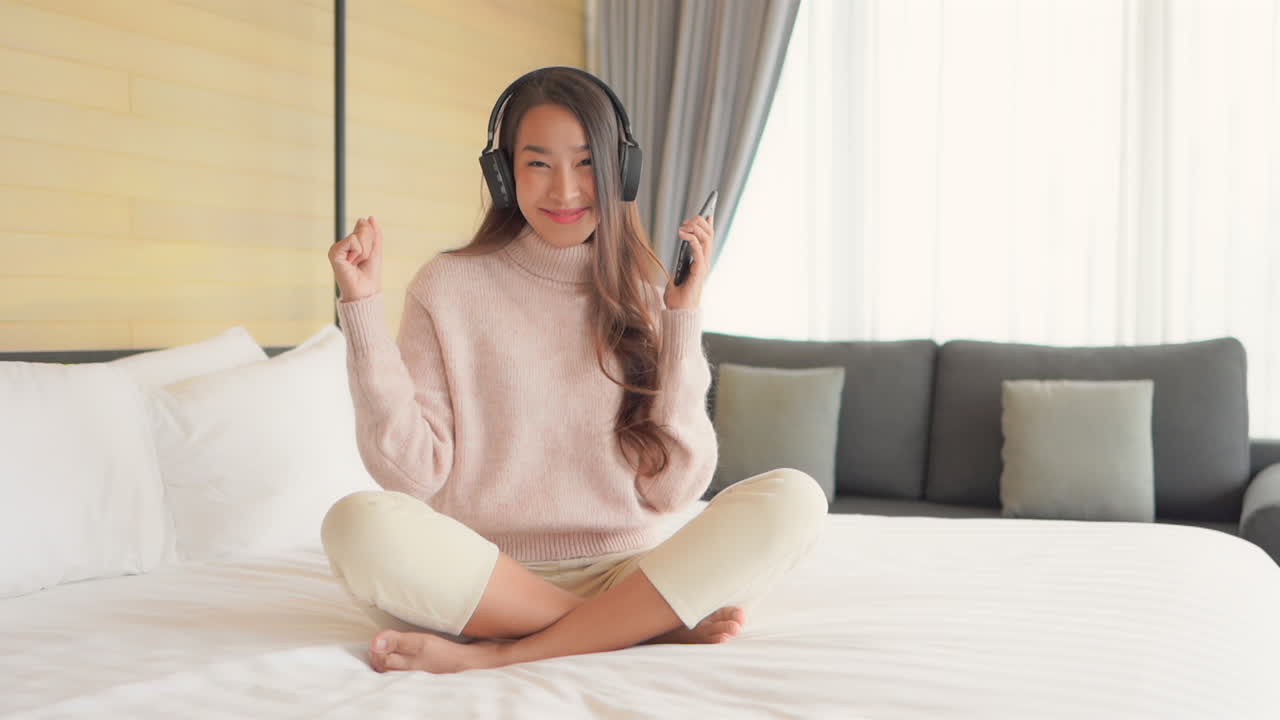 Pretty Asian girl grooving to dance music through bluetooth headphones while in bedroom. Fixed shot, Full shot