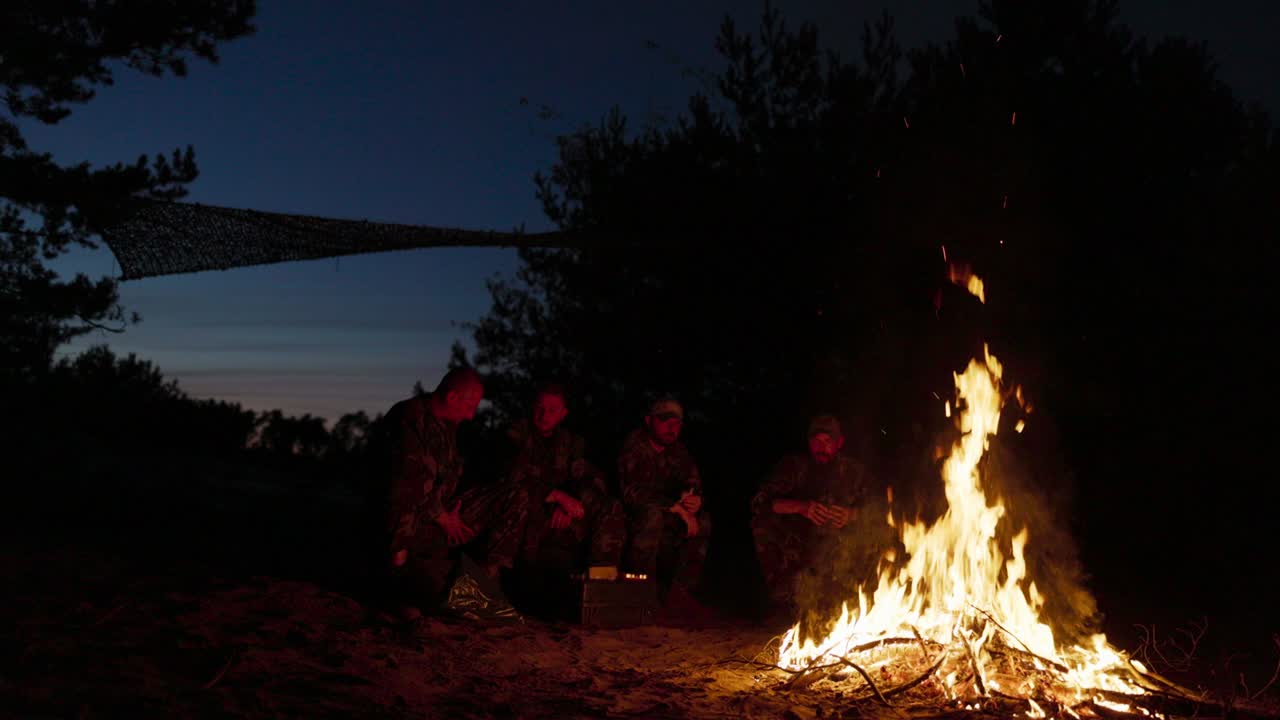 una gran llama de hoguera, detrás de ella hombres militares en uniformes de camuflaje están sentados, hablando y calentándose por la noche junto a la chimenea