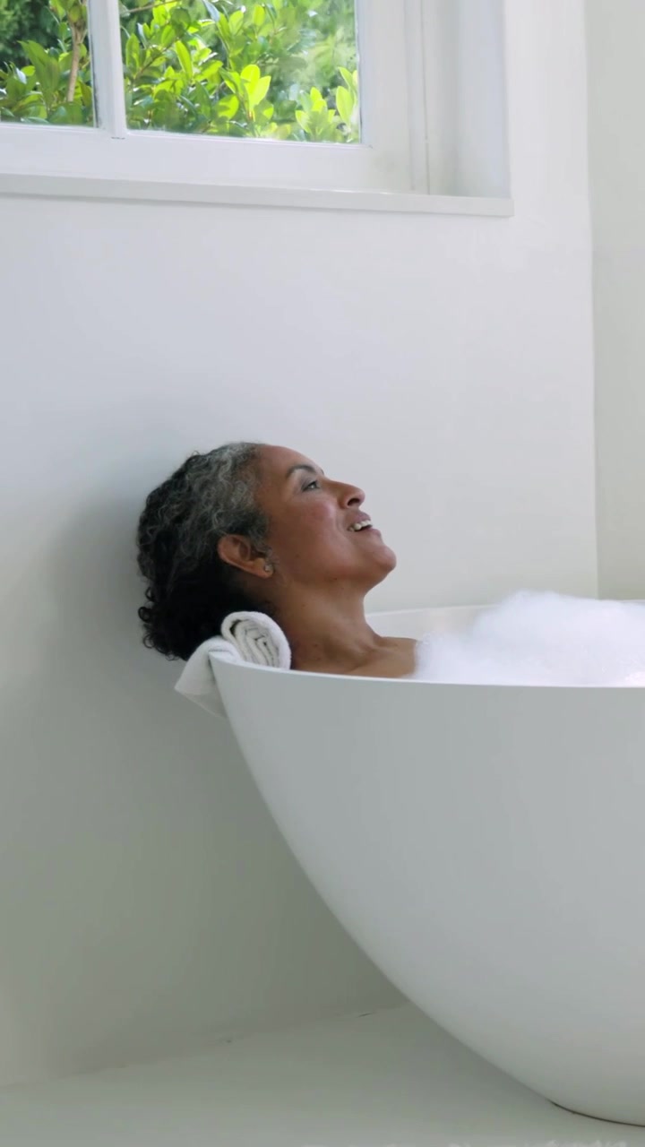 Vertical video: Camera closing in on woman relaxing in bathtub with rolled towel, foam, copy space
