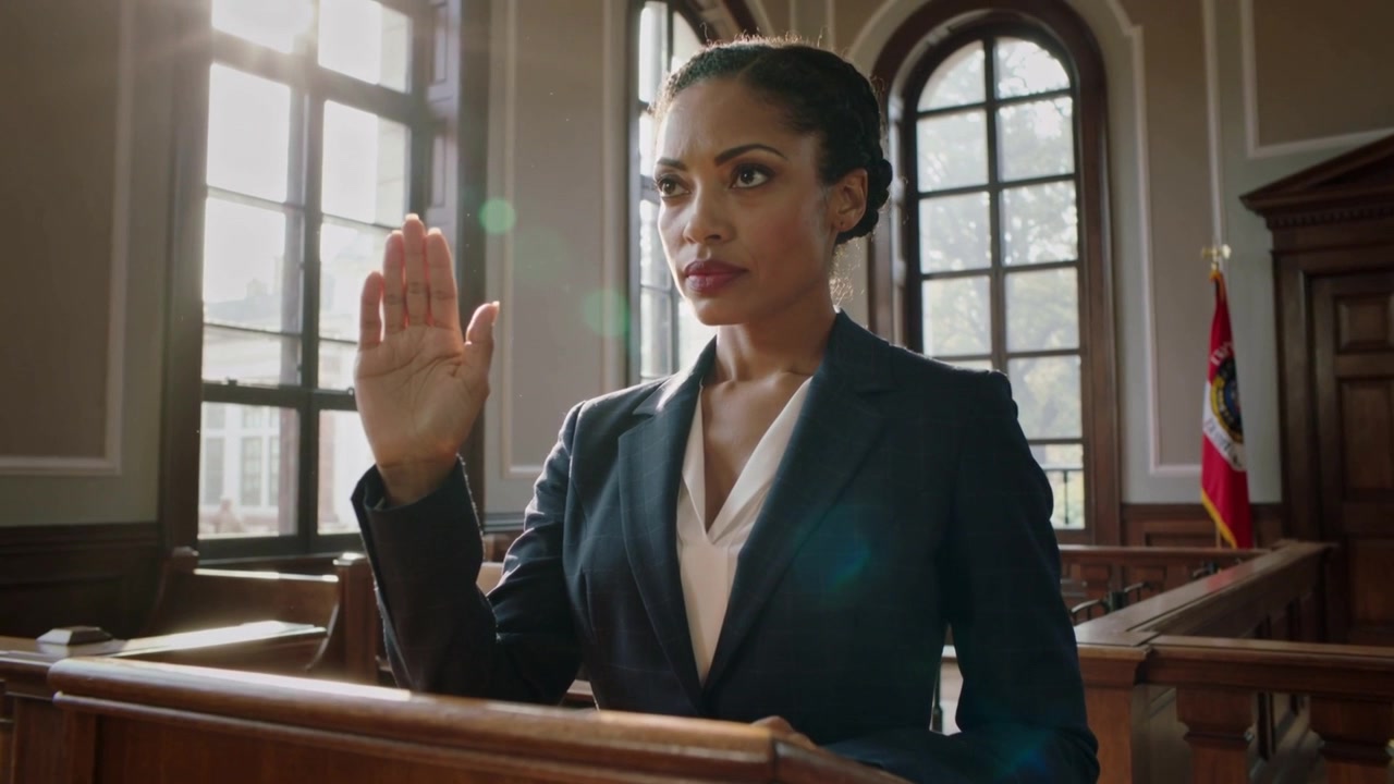 Donna in blazer blu scuro che risponde a una richiesta di giuramento, alzando la mano destra al leggio del tribunale, con bandiera visibile