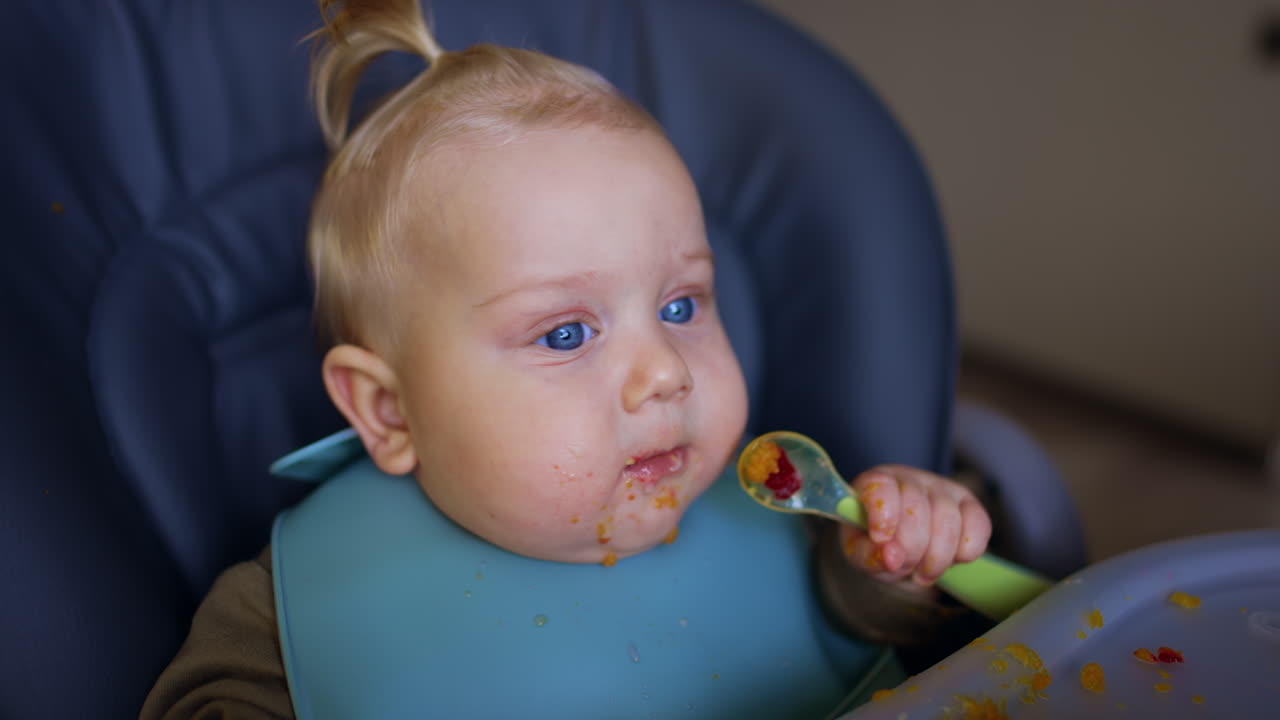 Little infant boy with smudged face sits at table. Cute kid taking a spoon from mom's hand.