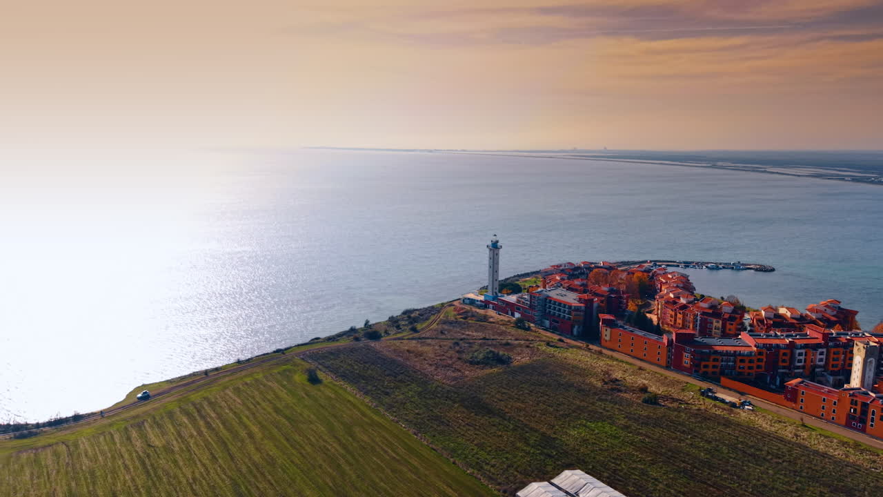 Filmato sul campo verde sulla costa del mare. Avvicinamento all'area residenziale colorata con un faro e un molo