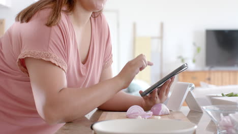 happy plus size biracial woman making meal with vegetables in kitchen and using tablet, slow motion