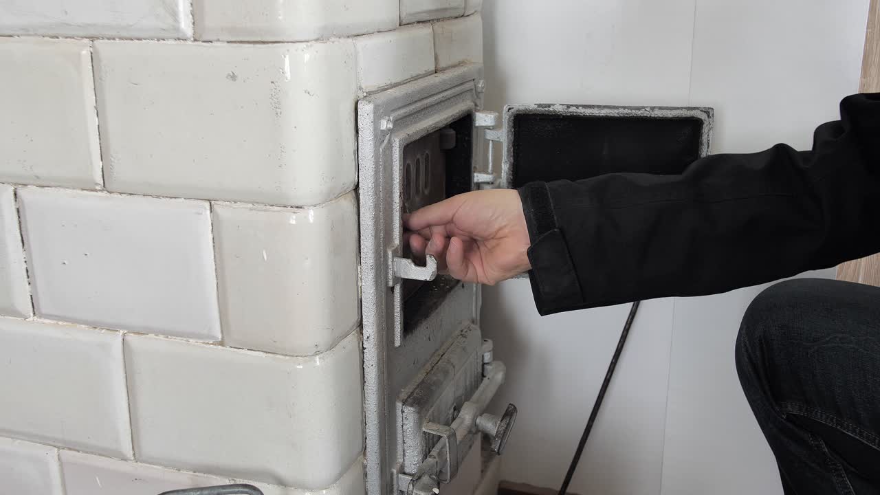 Man opens the door of an old home tiled stove and puts a firewood inside