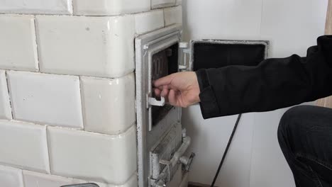 el hombre abre la puerta de una vieja estufa de azulejos y pone leña dentro