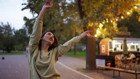 A-brunette-girl-dances-in-a-green-sweater-against-the-backdrop-of-a-cafe-in-the-park.-Fun,-holiday-atmosphere.-Happiness