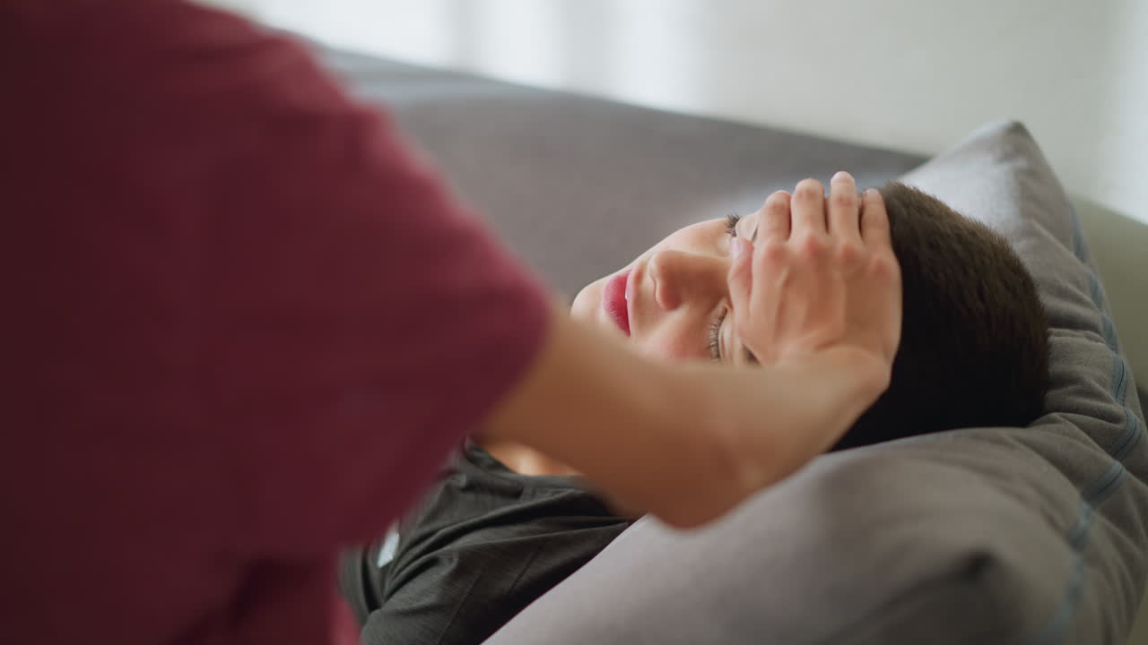 Close-up of someone placing hand on forehead of sick child lying on the couch, the child looks weak, and someone is observing with concern, offering care