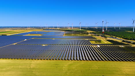 fields with solar panels in the countryside. multiple wind turbines rotate at backdrop. aerial view