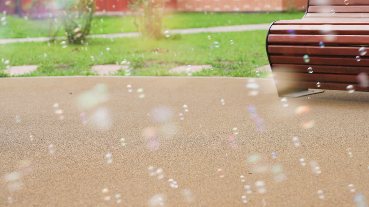 Close up of colorful soap bubbles drifting over pavement near bench, greenery and grass behind, gentle breeze carries tiny spheres across courtyard, soft bokeh sparkles