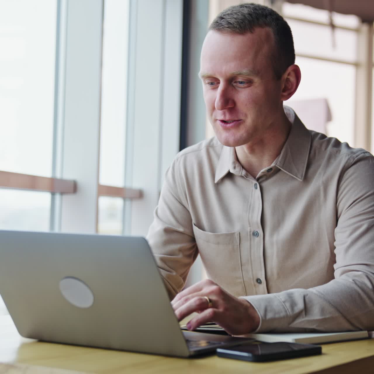 Smiling young man working at his laptop. Male employee says something and looks into window. Blurred backdrop