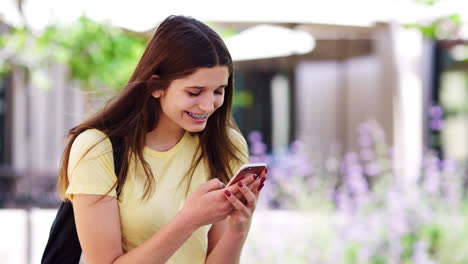 Female-High-School-Student-Checking-Messages-On-Mobile-Phone-Outside-College-Buildings