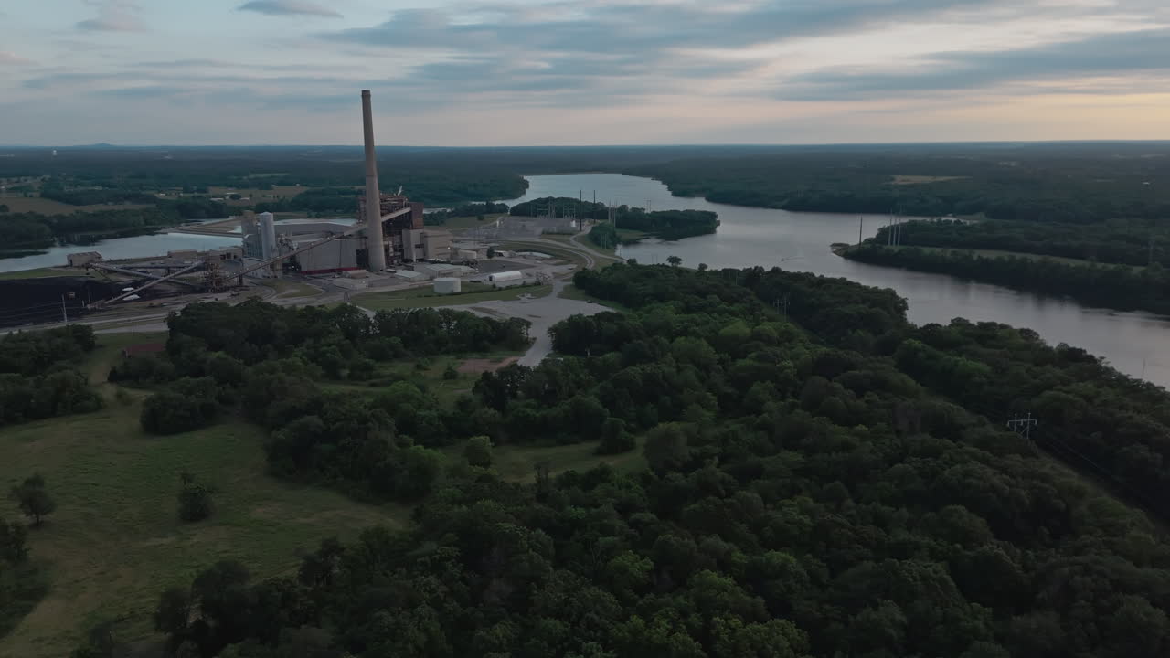 Drone ved kraftværk til industriel behandling langs bredden af en smal vandkanal, der adskiller skovområder med afdæmpet eftermiddagslys ovenover
