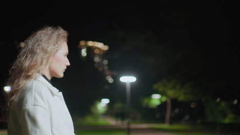 side view of young woman in white coat walking alone at night with thoughtful expression, blurred background featuring trees and glowing lights