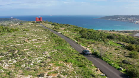 aerial drone view of a car driving on the paved road towards the red tower in mellieä§a, malta