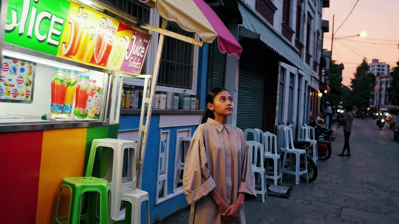 Young woman standing near a vibrant juice stand on a bustling city street at sunset, gazing upwards with a thoughtful expression, lost in contemplation as the golden hour light envelops her