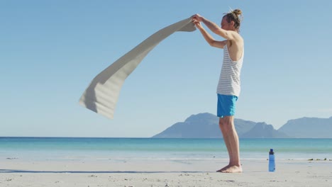 Video-Eines-Kaukasischen-Mannes-Mit-Dreadlocks,-Der-Am-Sonnigen-Strand-Eine-Yogamatte-Legt