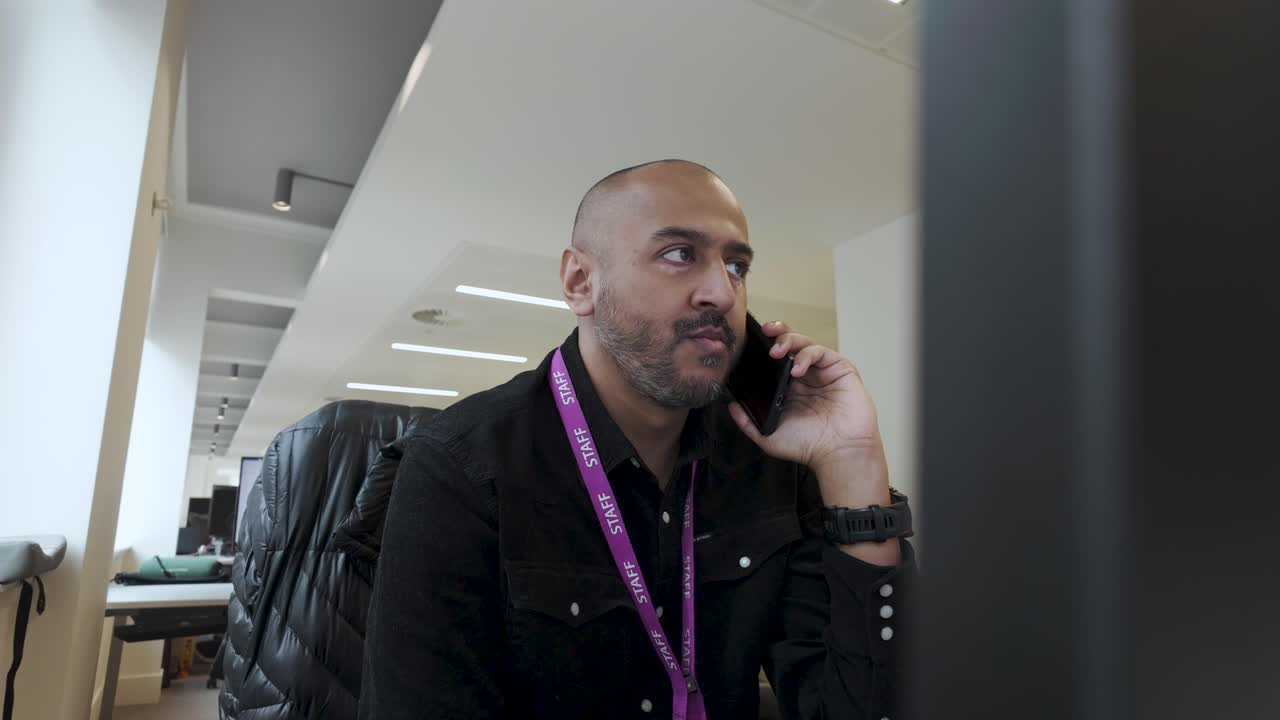 An individual is positioned indoors, facing the computer screen, making a phone call using a mobile phone