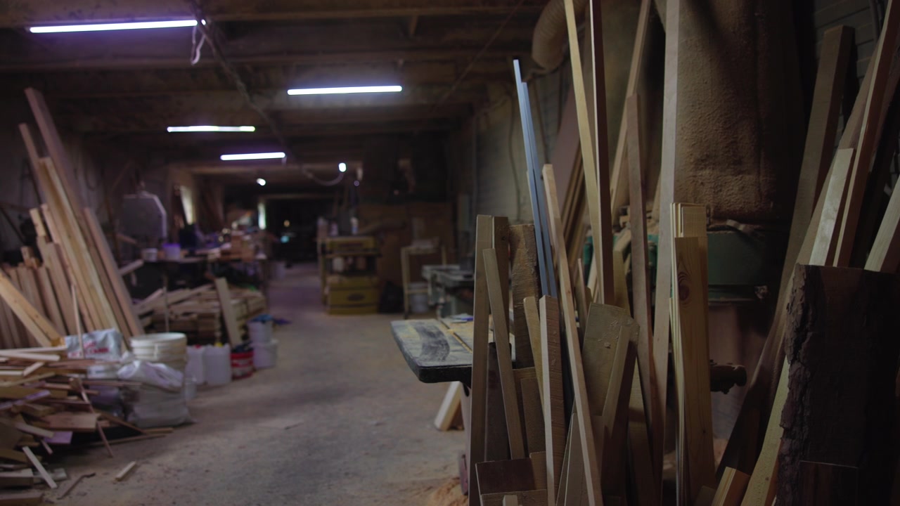 Stacked lumber beside workbench in narrow woodworking shop aisle under cool fluorescent lighting, with sawdust and wood scraps staged along long corridor
